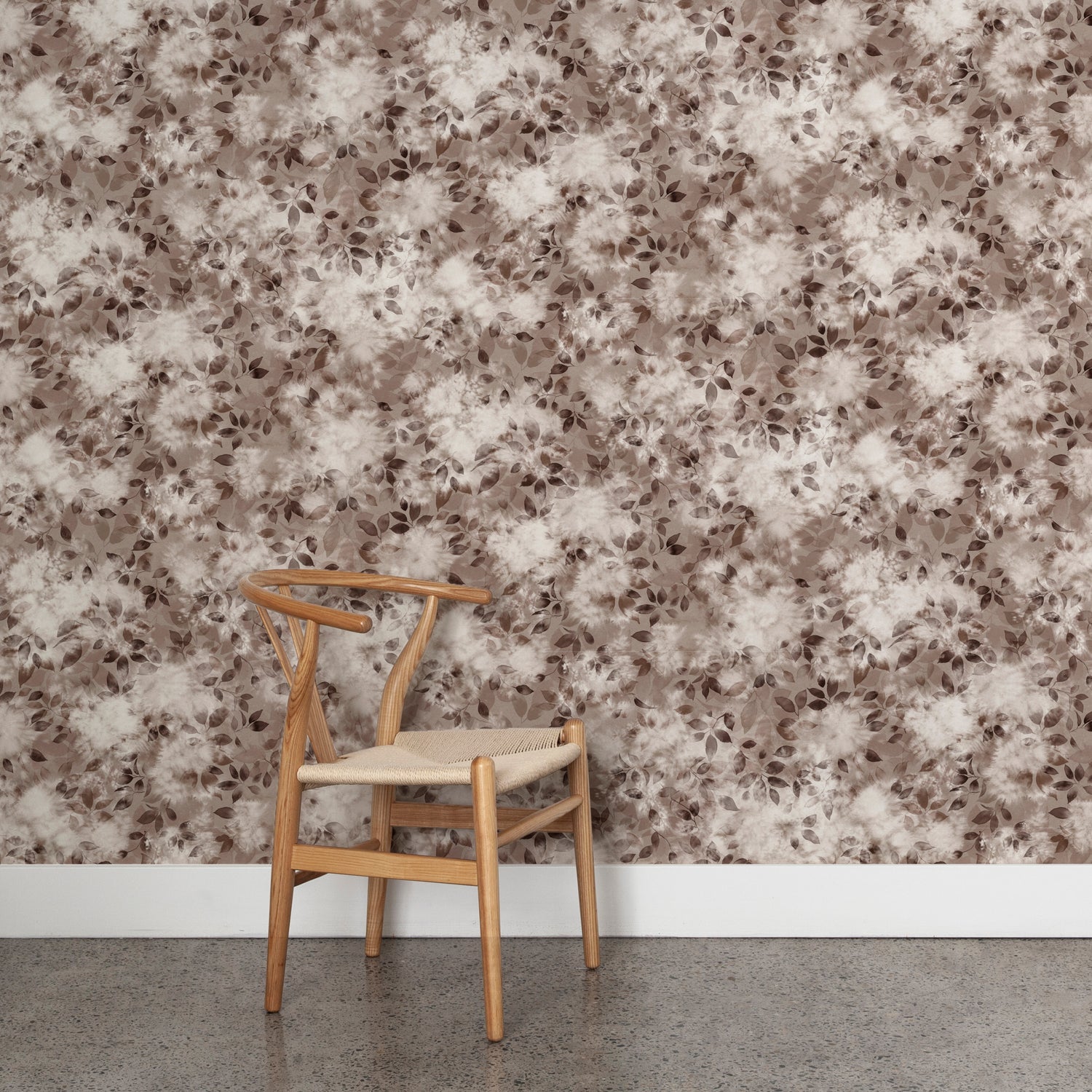 Angled wooden chair with woven seat on concrete floor against wallpaper in a painterly leaf print in shades of warm wine purple with a mottled white overlay.