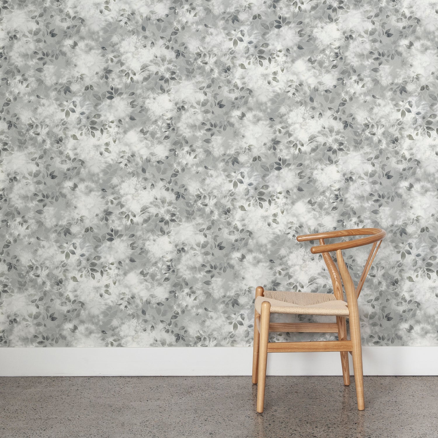Angled wooden chair with woven seat on concrete floor against wallpaper in a painterly leaf print in shades of cool steel grey with a mottled white overlay.