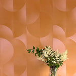 A vase of flowers stands in front of a wall covered in an abstract curvilinear print in mottled orange and pink.