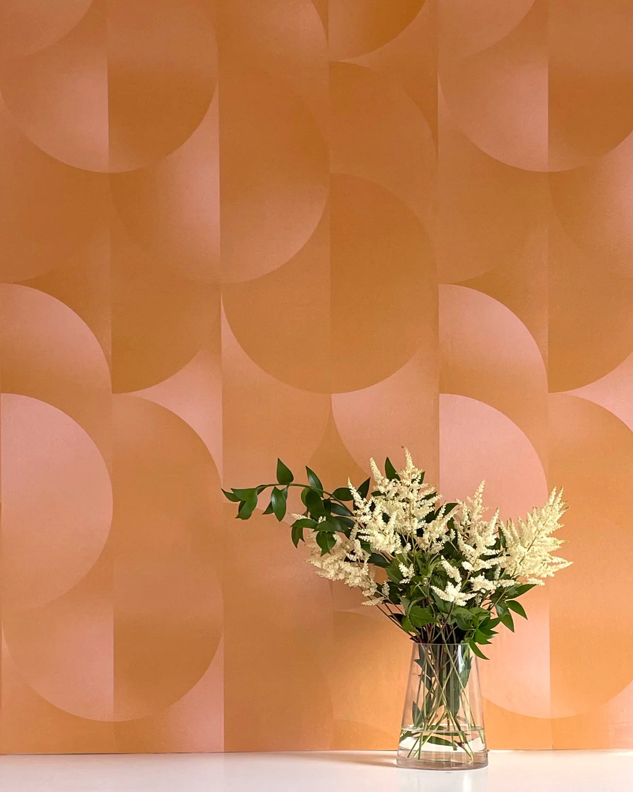 A vase of flowers stands in front of a wall covered in an abstract curvilinear print in mottled orange and pink.