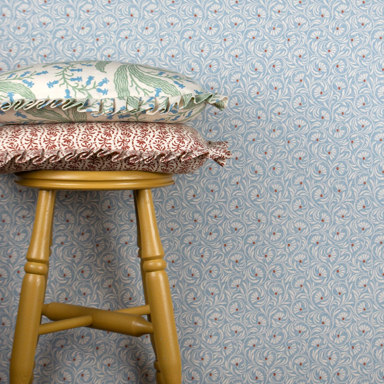 Wooden stool with decorative pillows against wallpaper in a lyrical wildflower design in white with red accents on a blue field. 