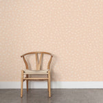 A wooden chair stands in front of a wall papered in a minimal repeating leaf print in cream on a light pink field.