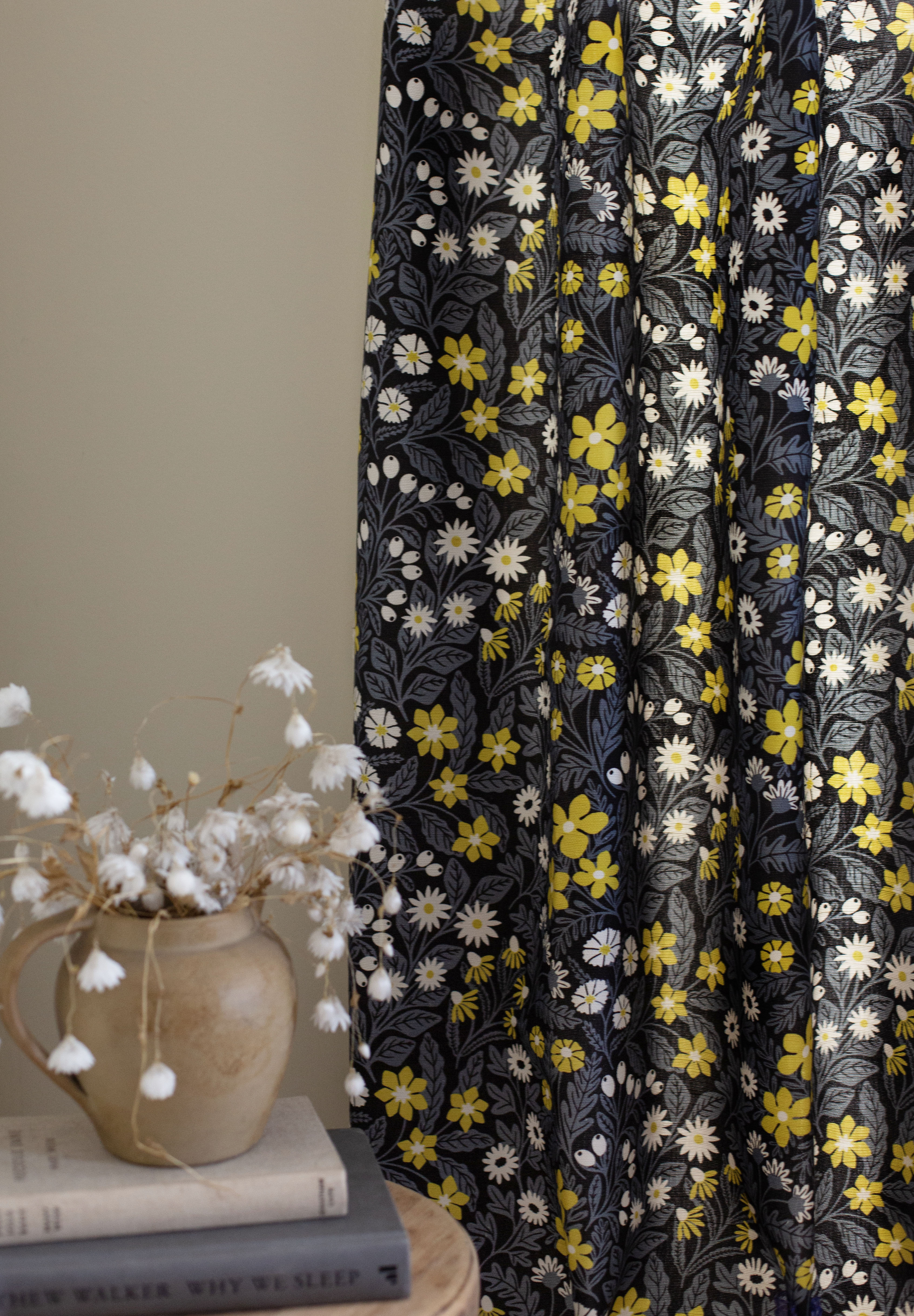 A vase of flowers stands in front of a draped curtain in a small-scale floral print in white, gray and yellow.