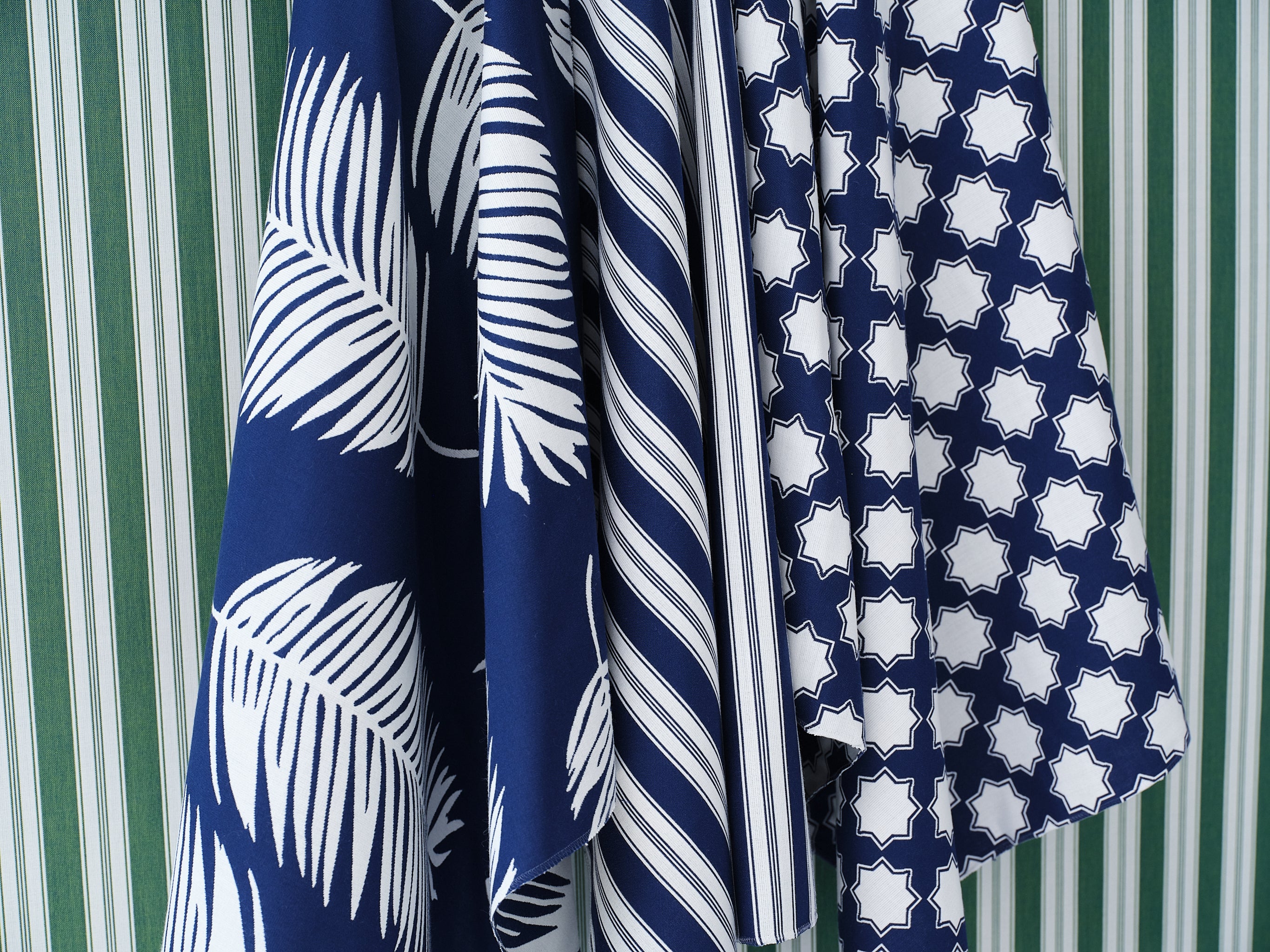 Hanging swatches of performance fabric in blue and white in a palm frond, stripe and moroccan star pattern on a green and white striped background.
