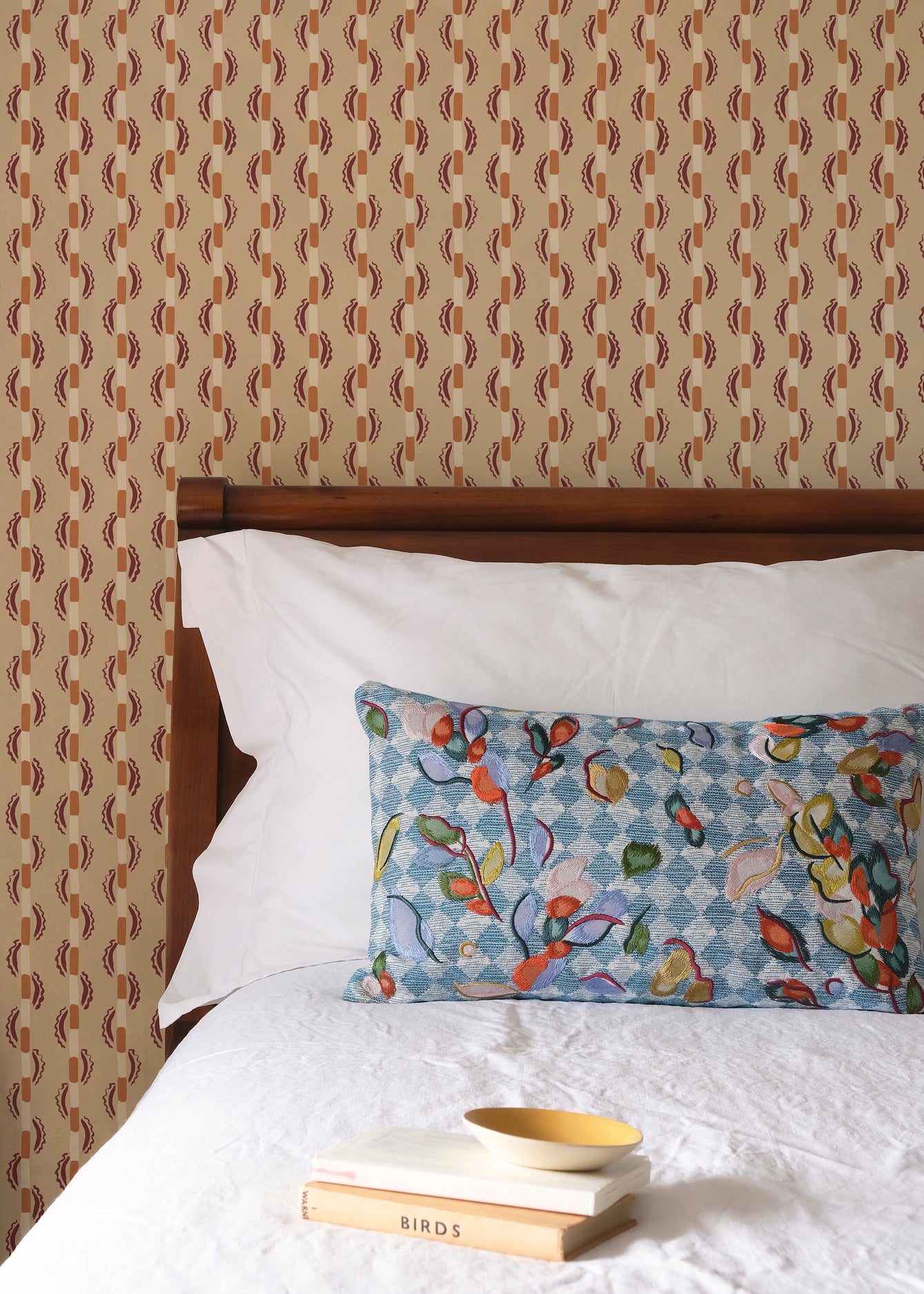Bed with patterned headboard, floral pillow, and books on a brown striped wallpaper.