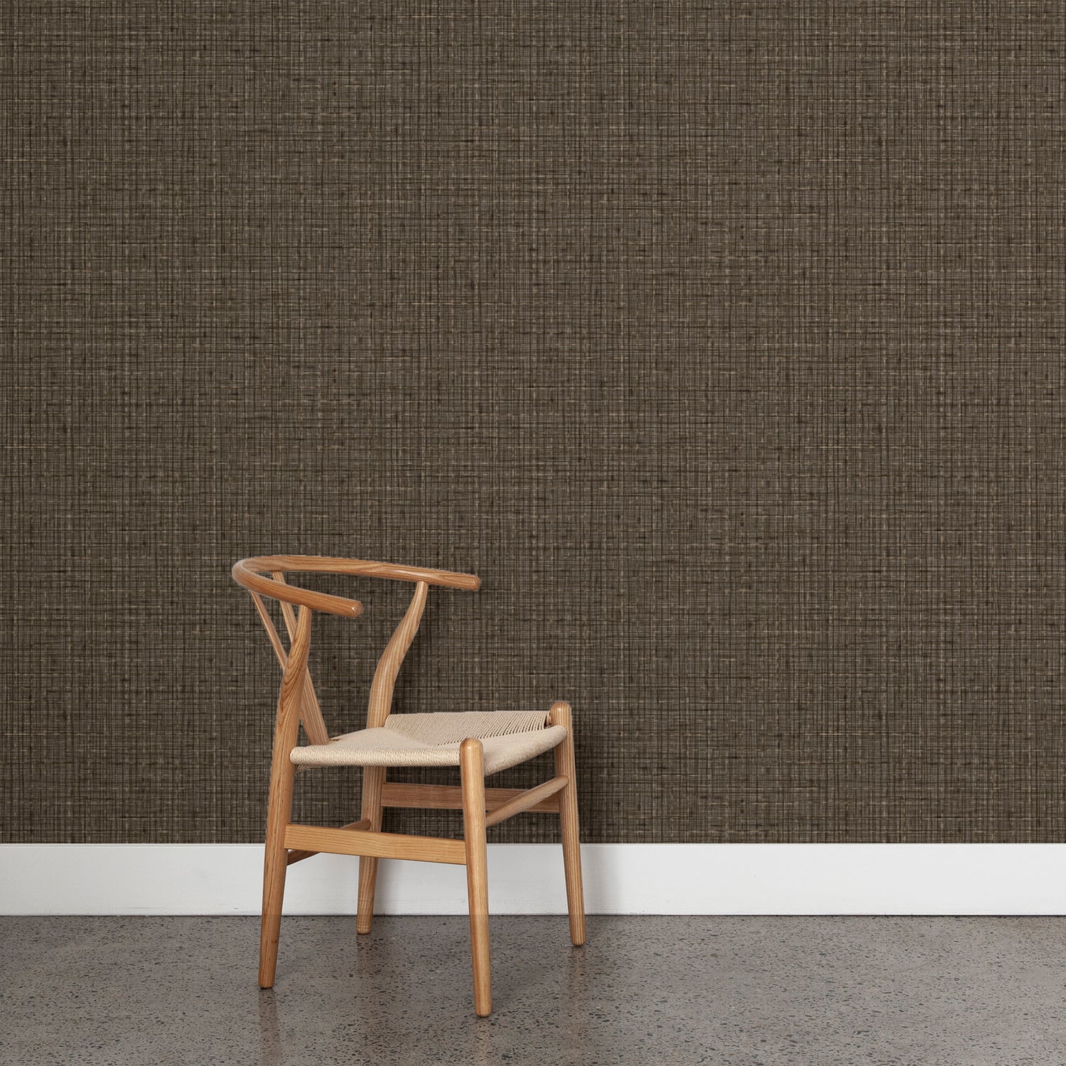 Angled wooden chair with woven seat on concrete floor against wallpaper in a textured tweed pattern in shades of warm charcoal grey.