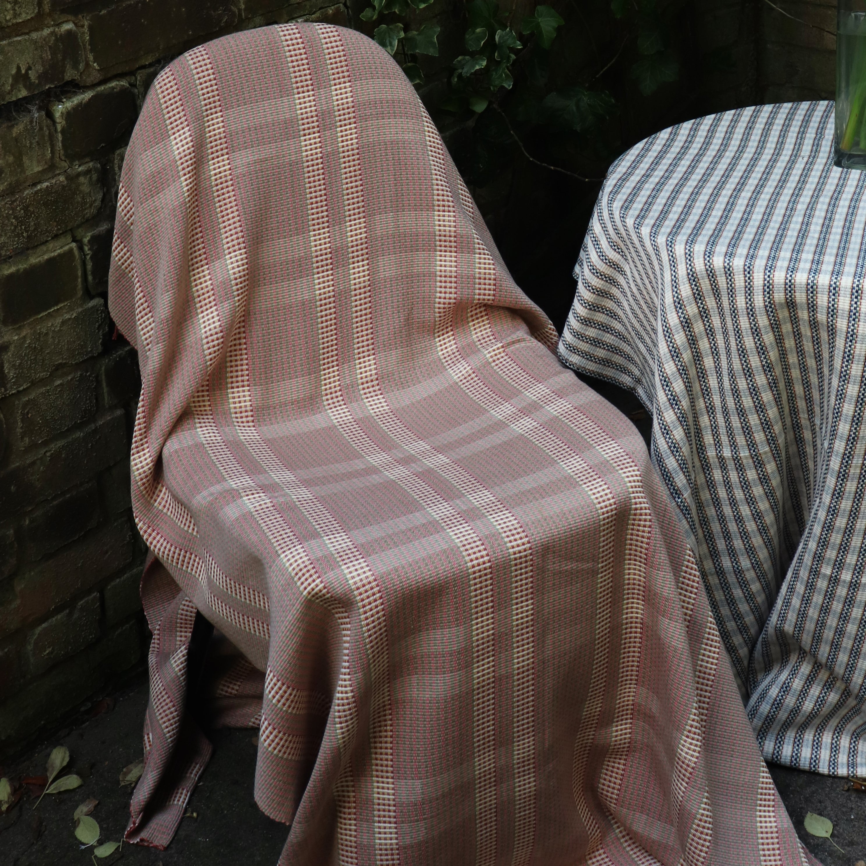 A courtyard outdoor vignette with a chair draped in a handwoven red window pane fabric, along side a table draped in a striped fabric.