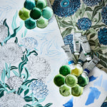 Overhead image of an artists process working on a botanical floral design n shades of blue and green with paint tubes, palettes and fabric background.