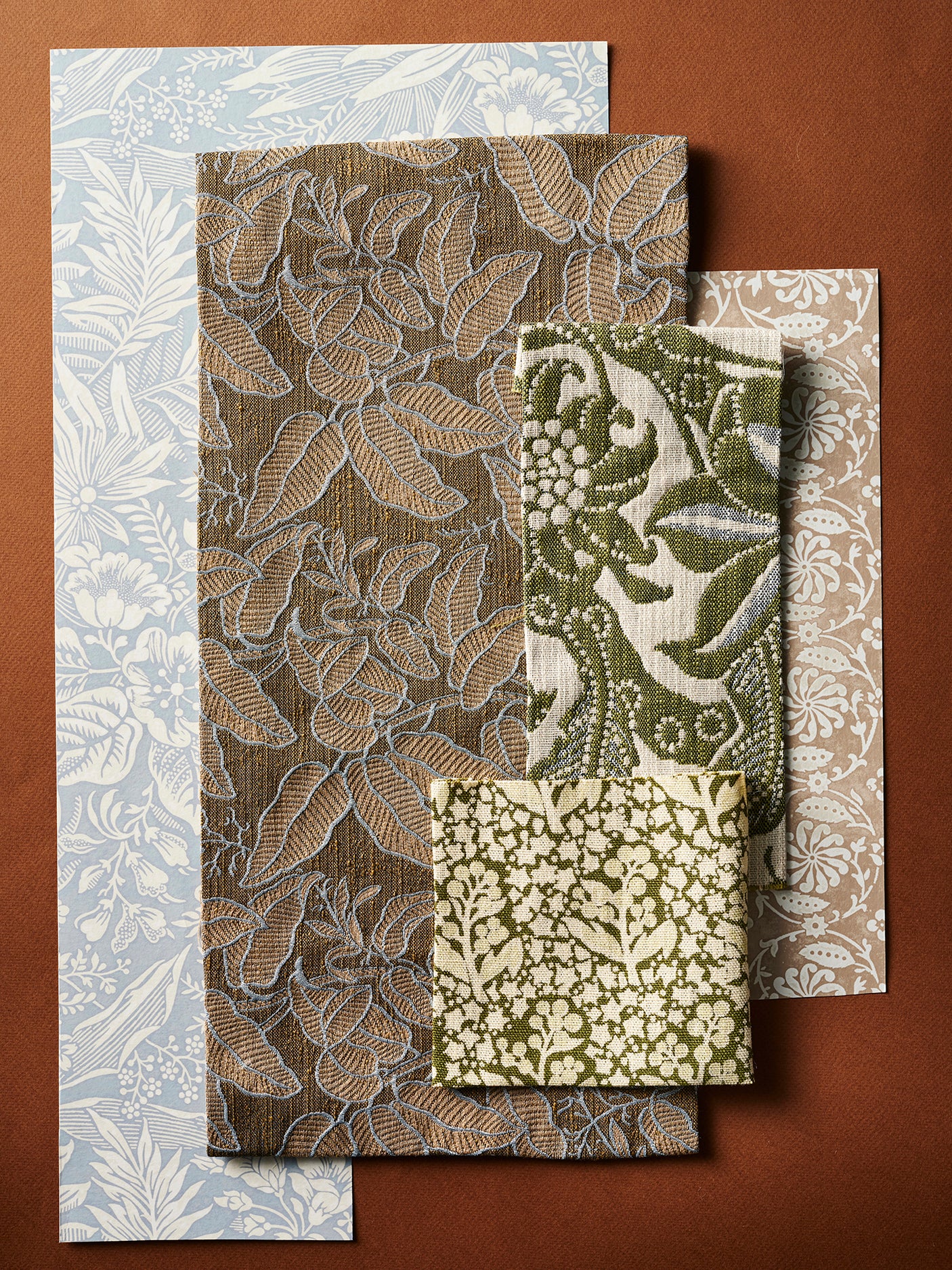 A flatlay with fabric and wallpaper swatches with botanical and floral patterns in muted tones of blue, brown, olive green, and beige, arranged on a solid brown background.