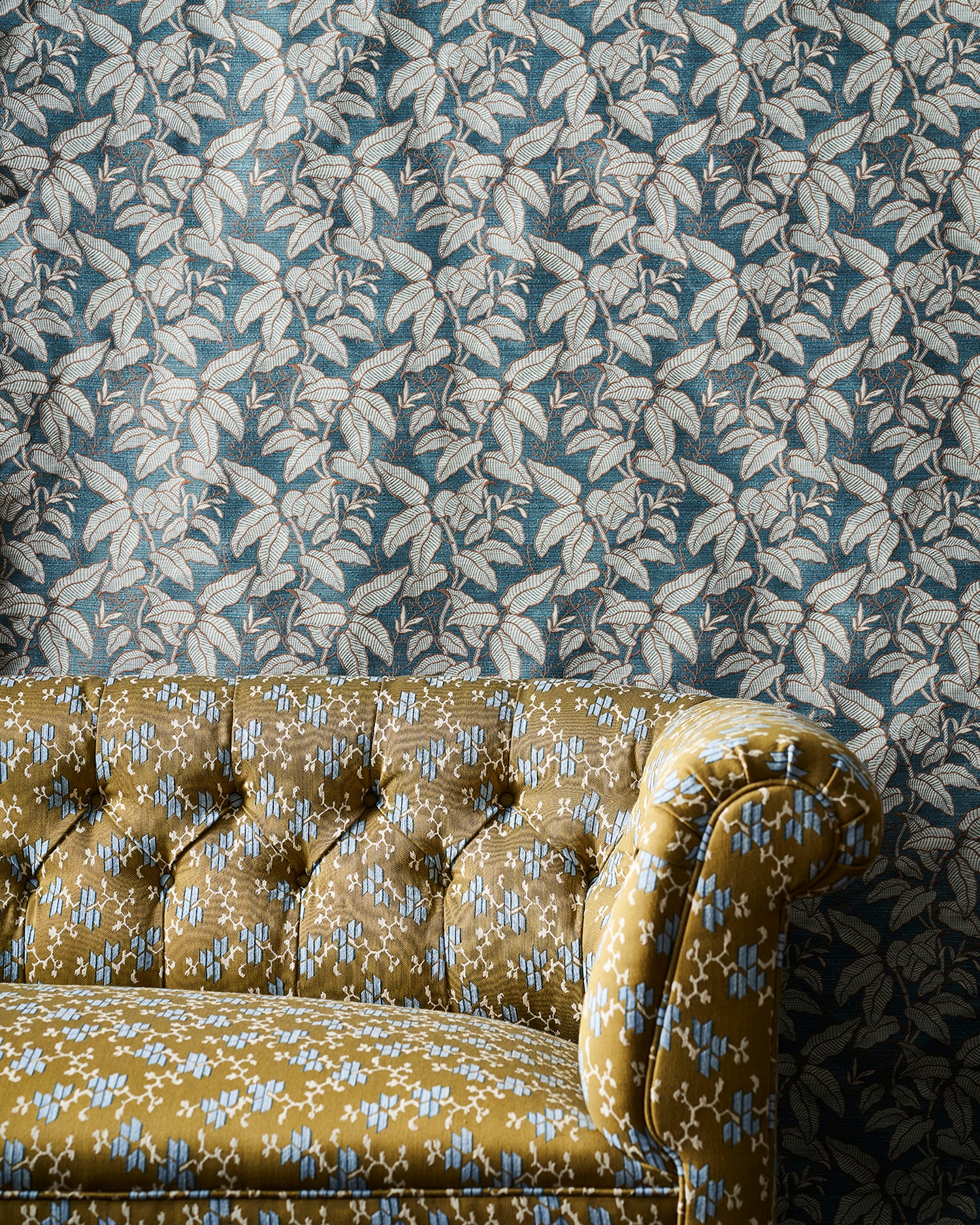 A close-up of a tufted couch upholstered in mustard brown fabric with a light blue floral pattern, set against a fabric  backdrop in a blue and white leaf motif