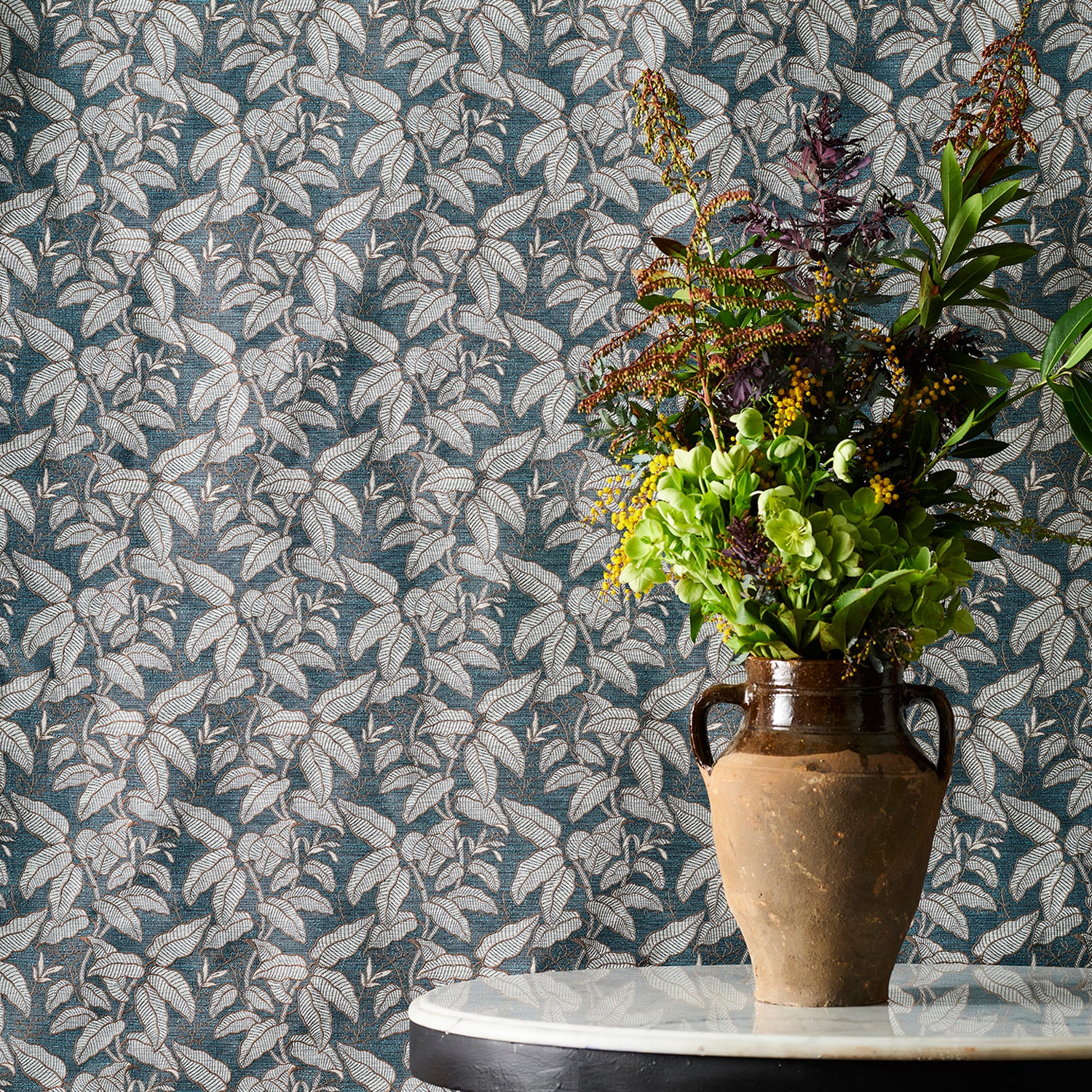 A white marble-topped table with a brown ceramic vase filled with a colorful assortment of foliage against a fabric backdrop with a botanical leaf pattern in grey and blue. 