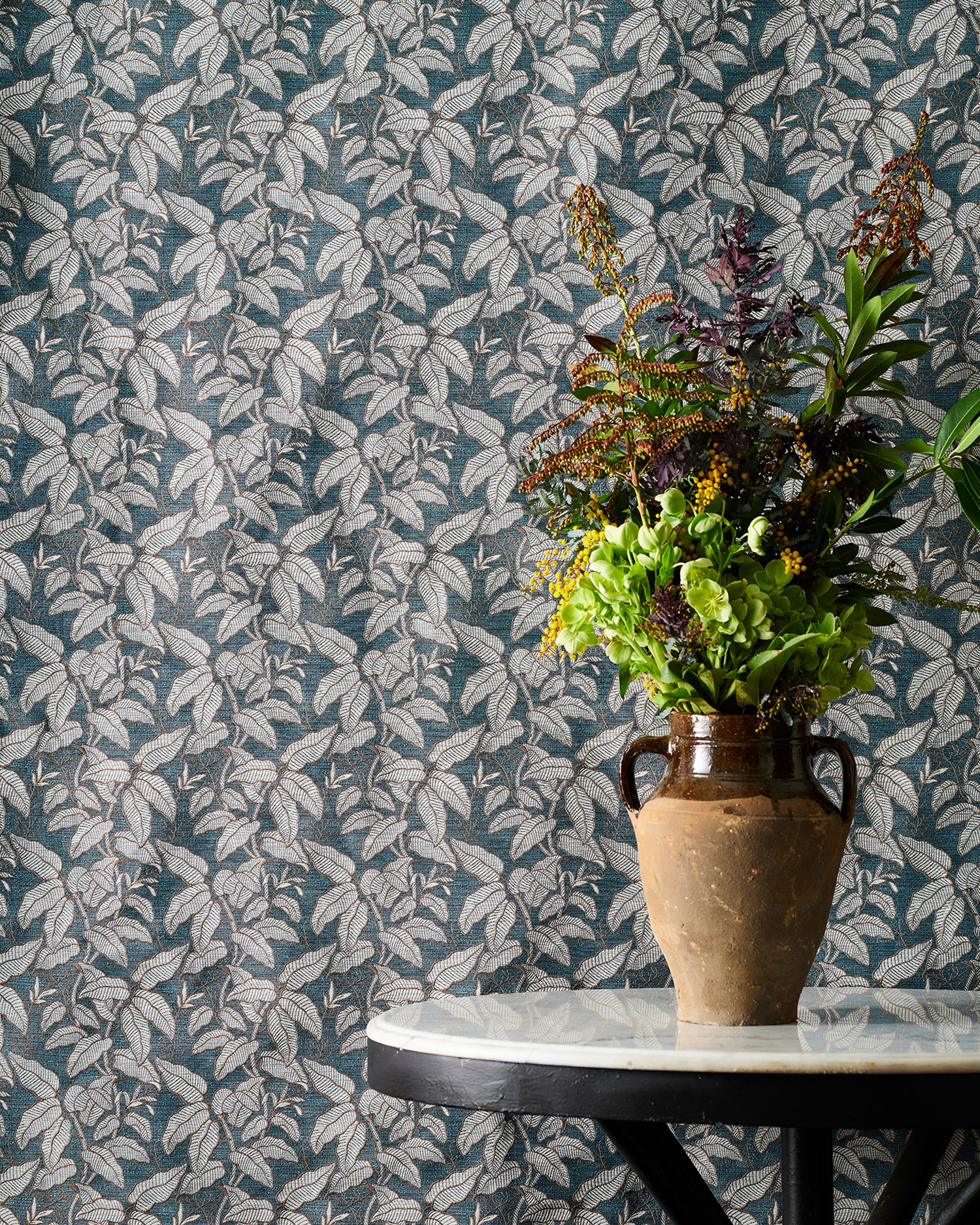 A white marble-topped table with a brown ceramic vase filled with a colorful assortment of foliage against a fabric backdrop with a botanical leaf pattern in grey and blue. 