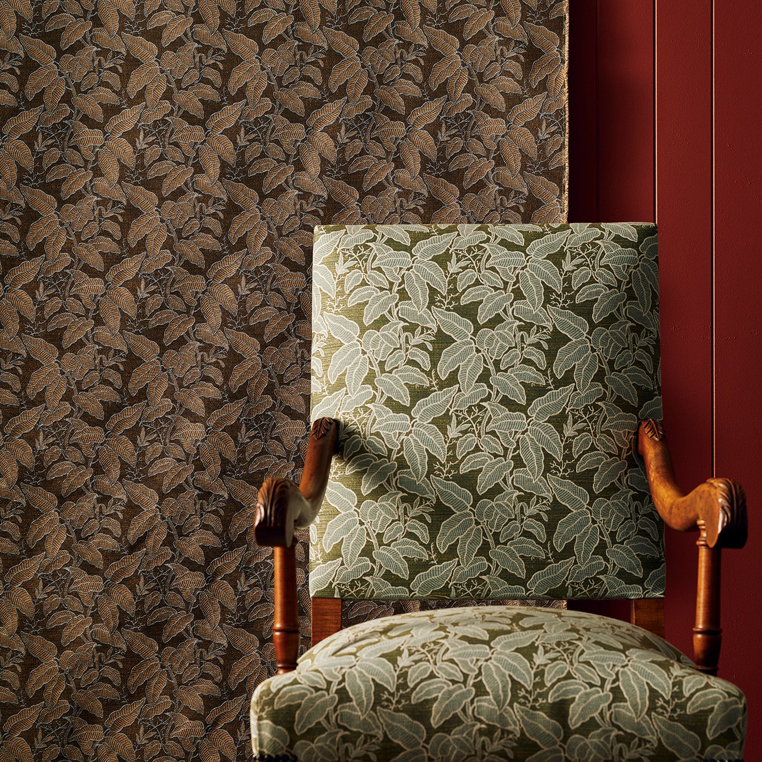 An upholstered wooden armchair with a leaf-patterned fabric in muted green and beige, set against a brown botanical fabric backdrop and a dark red wall panel.