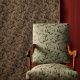 An upholstered wooden armchair with a leaf-patterned fabric in muted green and beige, set against a brown botanical fabric backdrop and a dark red wall panel.