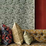 Three decorative throw pillows in floral and botanical patterns arranged on a tufted mustard-yellow sofa against a green leaf patterned fabric background.