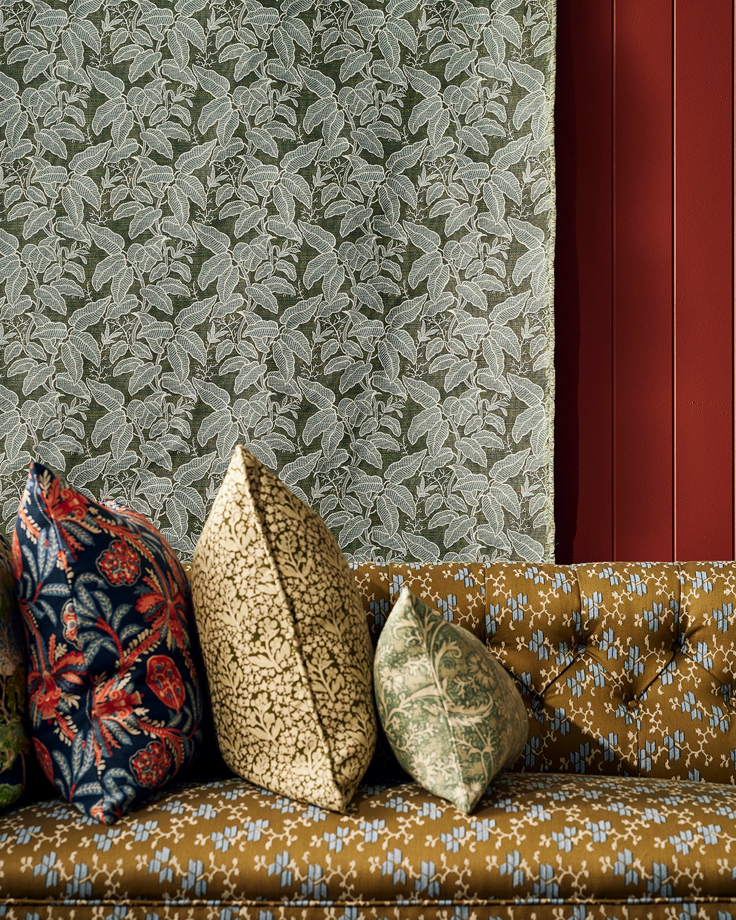 Three decorative throw pillows in floral and botanical patterns arranged on a tufted mustard-yellow sofa against a green leaf patterned fabric background.