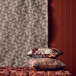 A styled interior scene showing patterned home textiles: a leafy curtain in taupe and brown, two decorative pillows with floral and ornamental prints, and a tufted upholstered bench in deep reddish-brown against a dark red paneled wall.