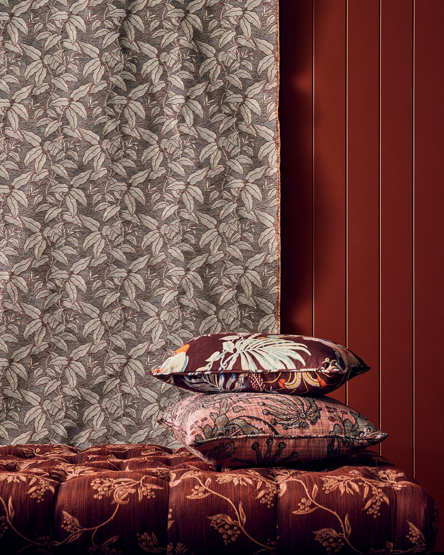 A styled interior scene showing patterned home textiles: a leafy curtain in taupe and brown, two decorative pillows with floral and ornamental prints, and a tufted upholstered bench in deep reddish-brown against a dark red paneled wall.