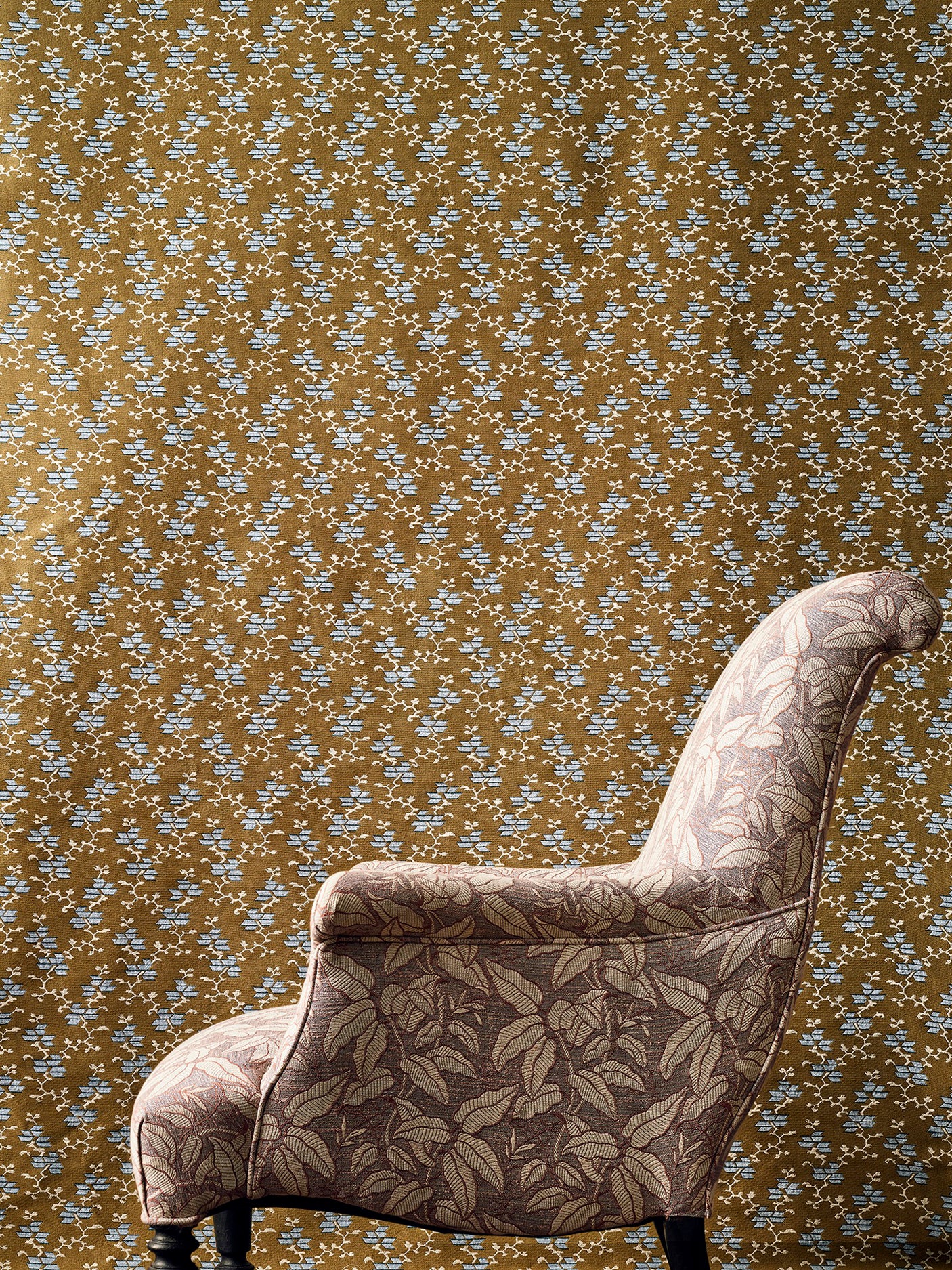 An upholstered armchair with a leafy floral pattern in beige and brown, set against a brown and grey small scale geo-floral fabric backdrop.