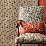 An antique-style wooden armchair upholstered in a blue and beige botanical patterned fabric, styled with a red floral throw pillow, set against small scale geo-floral fabric backdrop and a red wall panel.