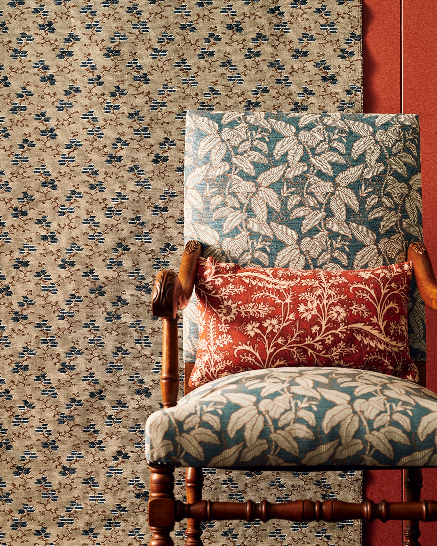 An antique-style wooden armchair upholstered in a blue and beige botanical patterned fabric, styled with a red floral throw pillow, set against small scale geo-floral fabric backdrop and a red wall panel.