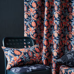 A close-up of botanical patterned fabric in a room setting, showing a curtain and matching cushion cover on a dark teal chair against a dark wall.