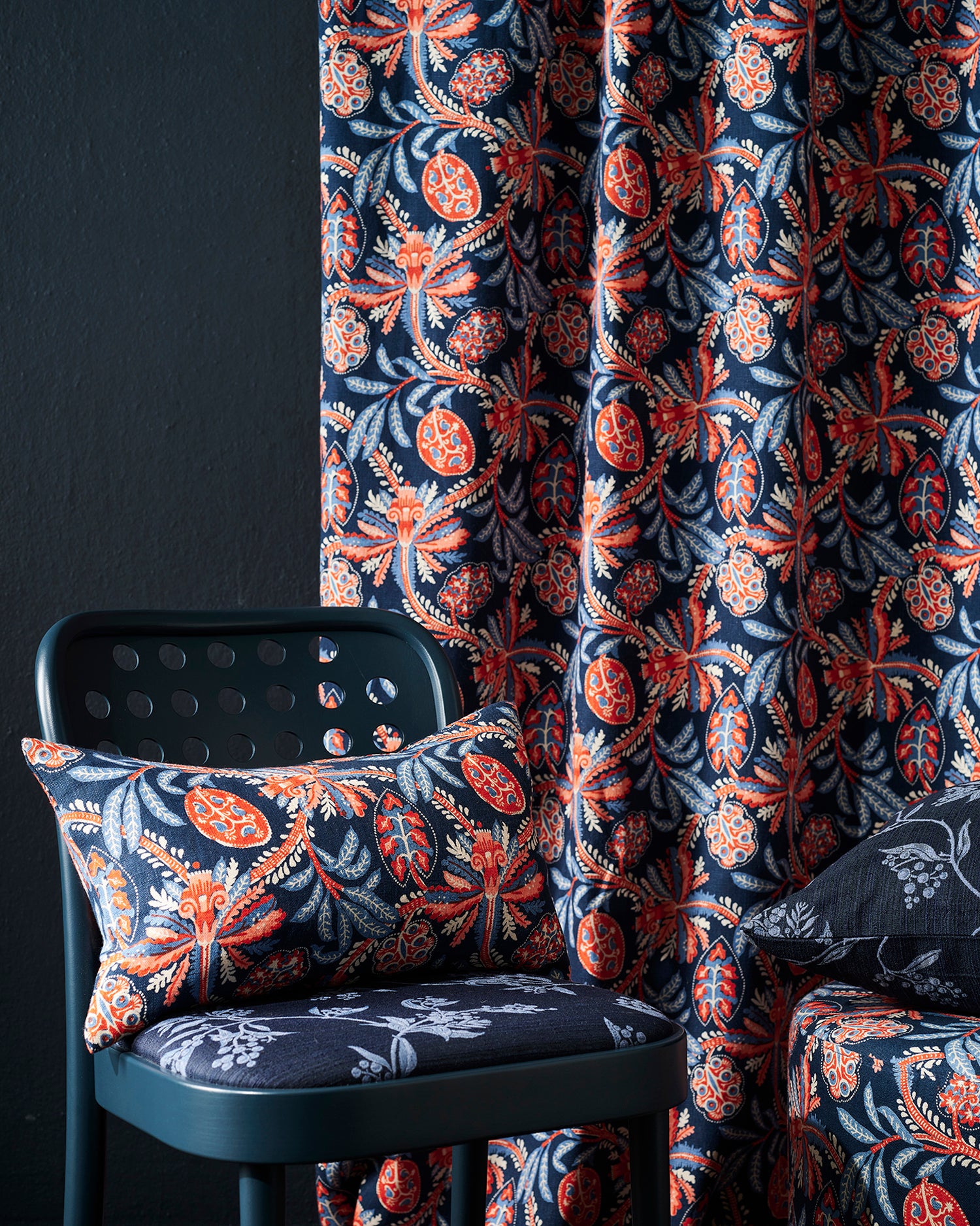A close-up of botanical patterned fabric in a room setting, showing a curtain and matching cushion cover on a dark teal chair against a dark wall.