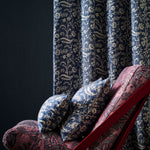 A close-up of botanical patterned curtains in navy blue and beige, with matching decorative pillows and a red-framed lounge chair in a dark room.