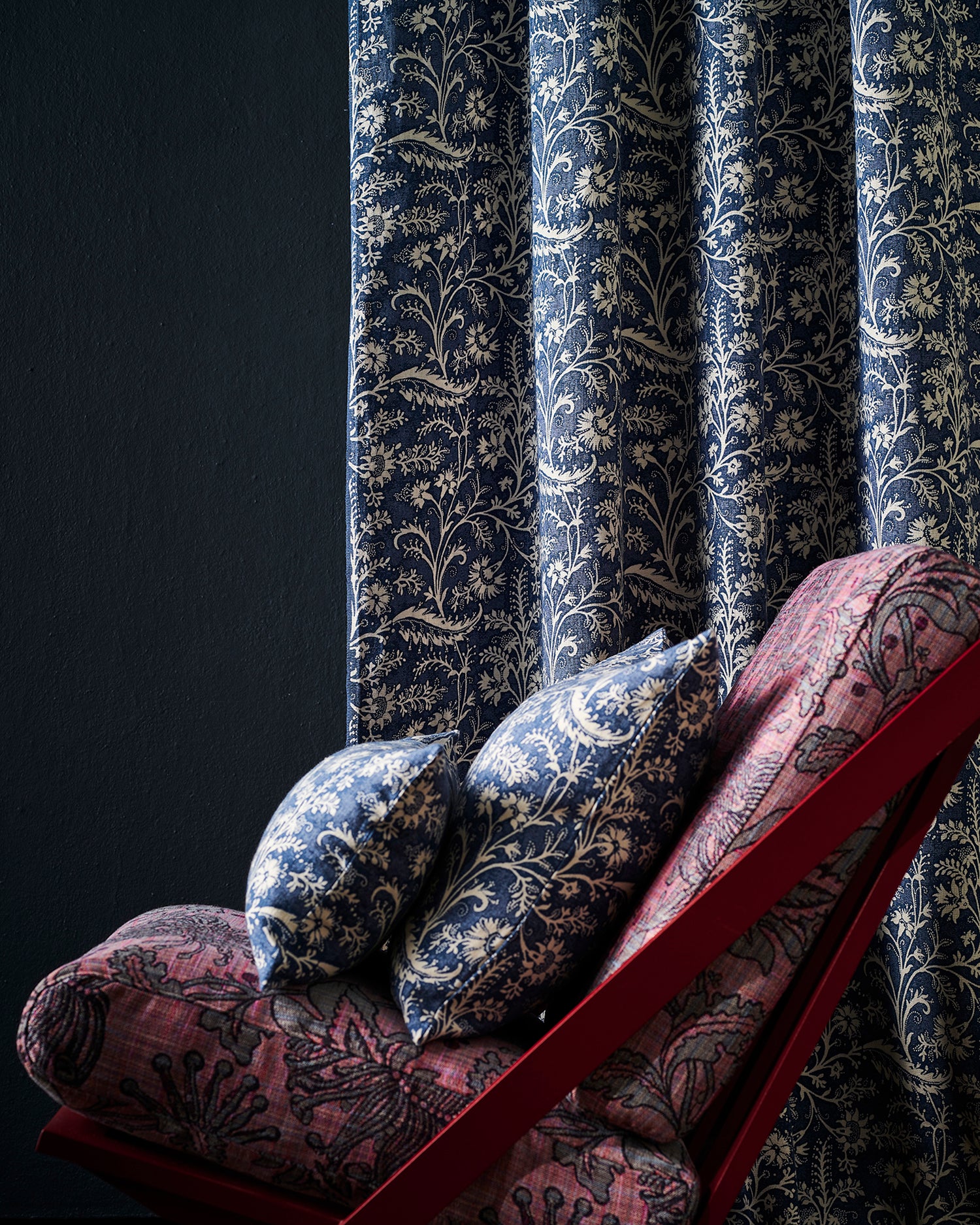 A close-up of botanical patterned curtains in navy blue and beige, with matching decorative pillows and a red-framed lounge chair in a dark room.