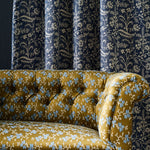 A close-up of a tufted velvet sofa upholstered in mustard yellow fabric with a light blue and white geo-floral pattern, set against a background of dark navy blue curtains with a cream botanical print.