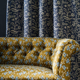 A close-up of a tufted velvet sofa upholstered in mustard yellow fabric with a light blue and white geo-floral pattern, set against a background of dark navy blue curtains with a cream botanical print.