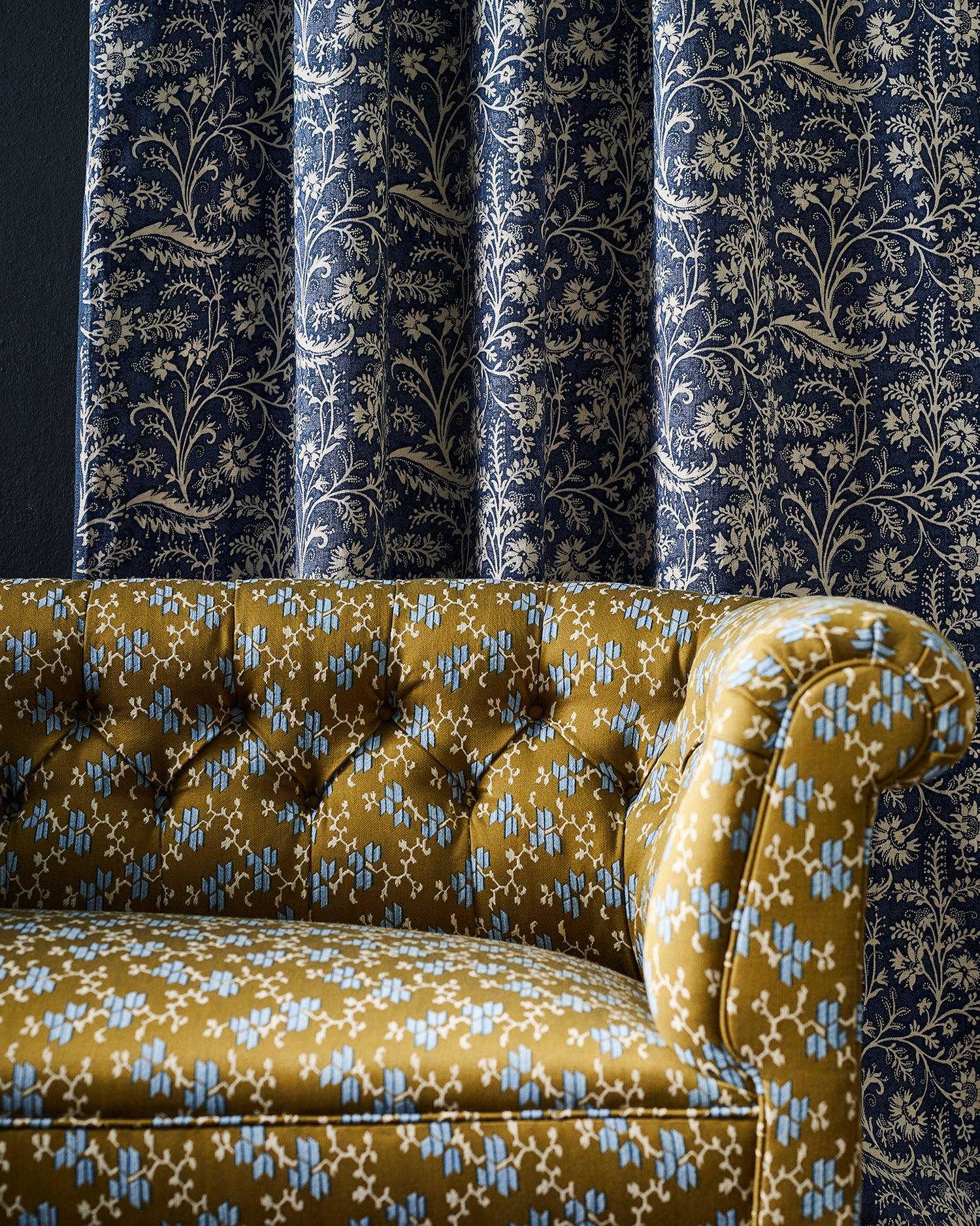 A close-up of a tufted velvet sofa upholstered in mustard yellow fabric with a light blue and white geo-floral pattern, set against a background of dark navy blue curtains with a cream botanical print.