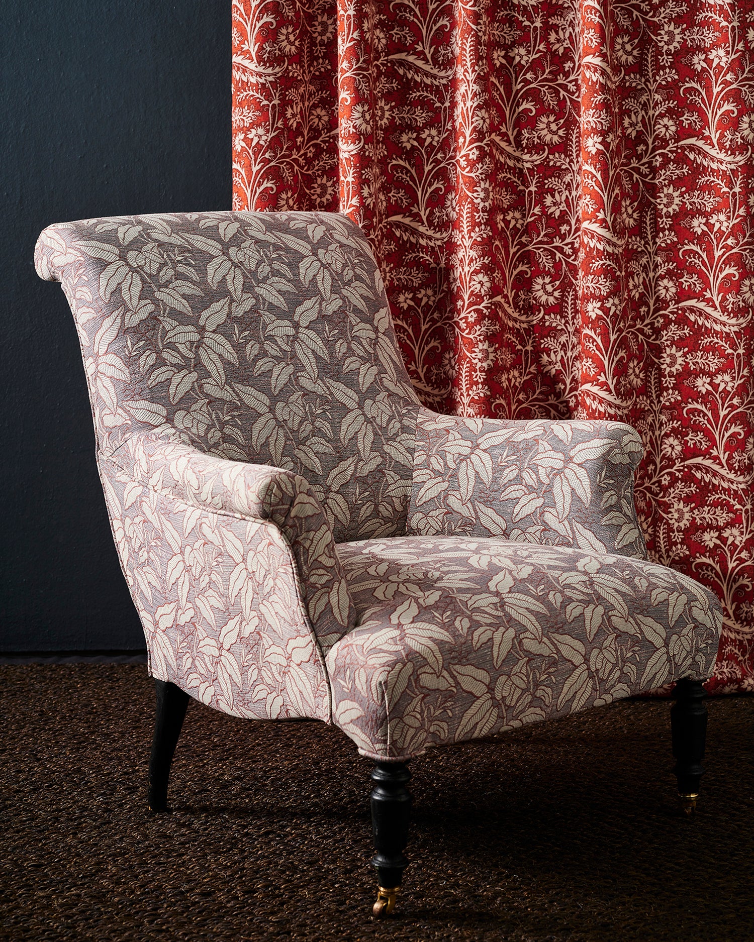A classic wingback armchair upholstered in a warm grey and cream botanical leaf patterned fabric, set against a dark blue wall and a red floral patterned curtain.