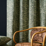 A close-up of a room featuring a green and beige botanical patterned curtain, a matching patterned cushion, and a wooden armchair with brown patterned upholstery.