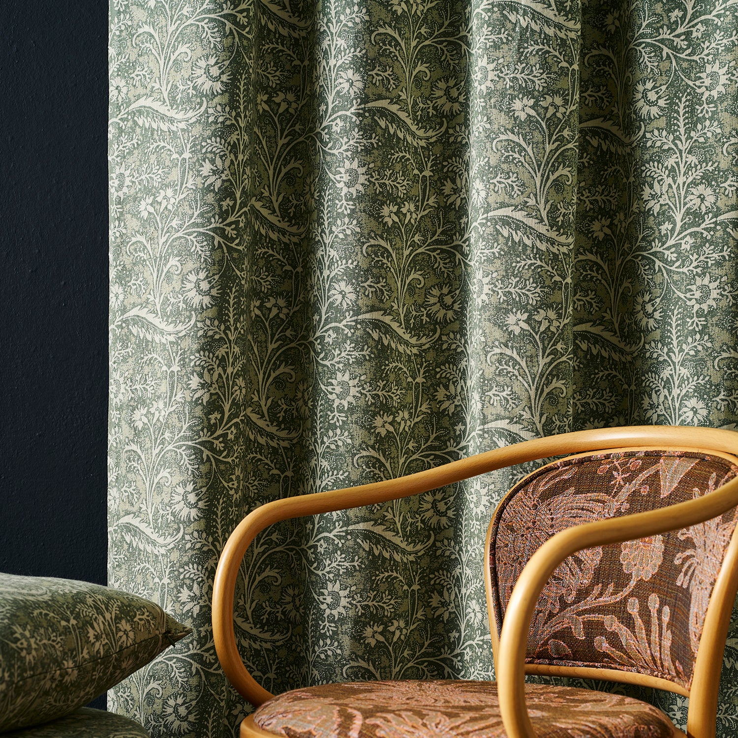 A close-up of a room featuring a green and beige botanical patterned curtain, a matching patterned cushion, and a wooden armchair with brown patterned upholstery.