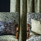 Two upholstered armchairs with botanical fabric in green, blue, and navy tones, arranged around a green floral vine patterned curtain panel against a dark background.