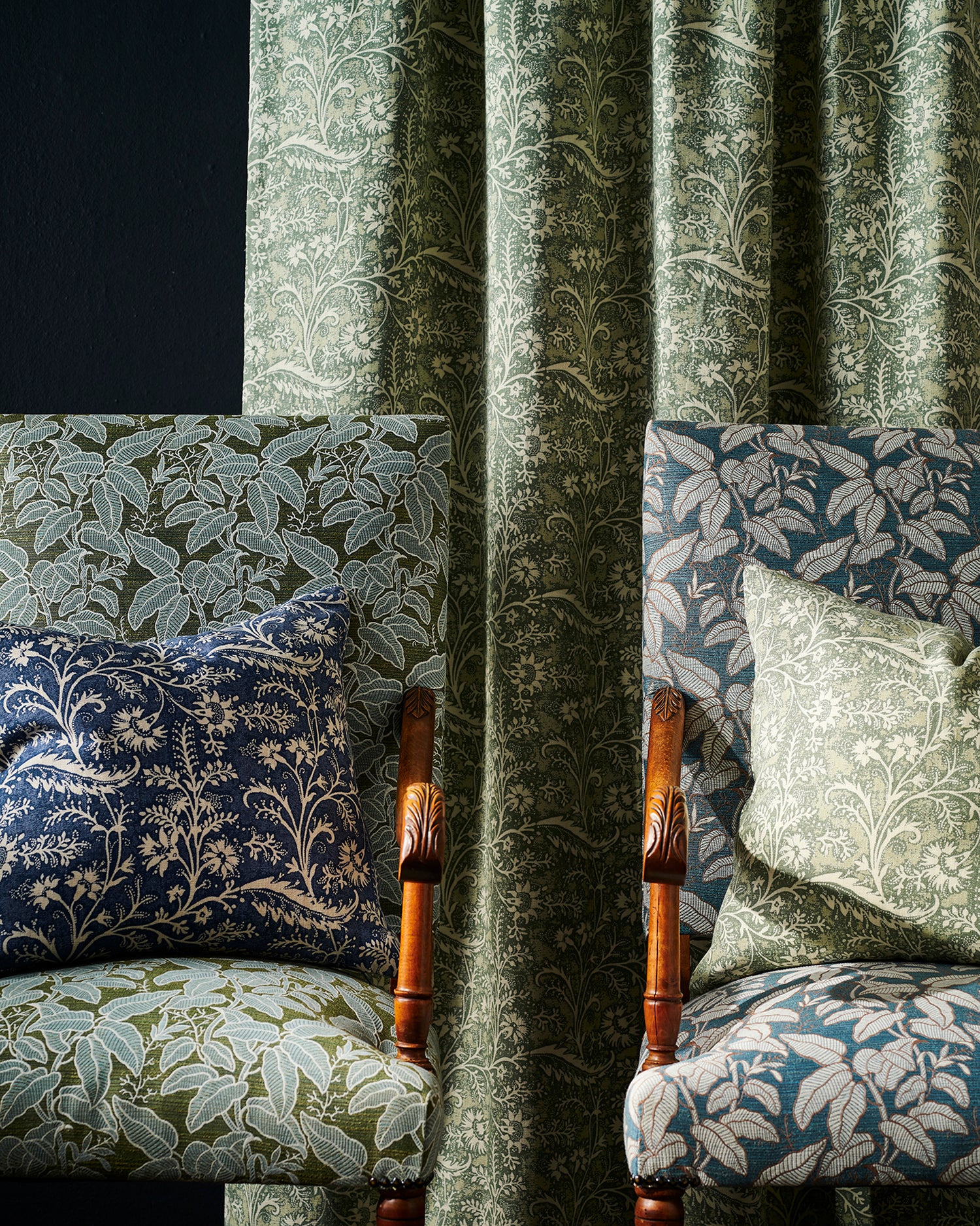 Two upholstered armchairs with botanical fabric in green, blue, and navy tones, arranged around a green floral vine patterned curtain panel against a dark background.