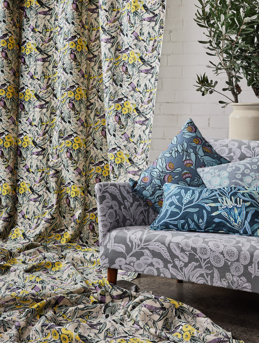 Vignette of a draped bird printed fabrc, with a grey floral patterned couch with a pile of botanical printed pillows. 