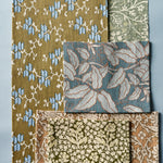 A set of woven fabric swatches with various botanical and geometric patterns, including olive green, dusty blue, and beige designs, arranged on a cool blue background.