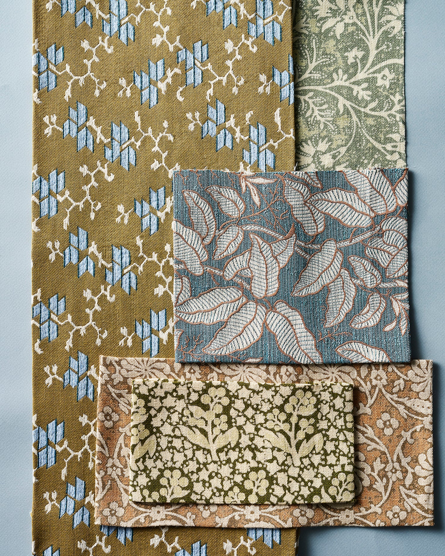 A set of woven fabric swatches with various botanical and geometric patterns, including olive green, dusty blue, and beige designs, arranged on a cool blue background.