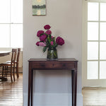 Floral arrangement in a vase on a wooden side table in a room with large windows.