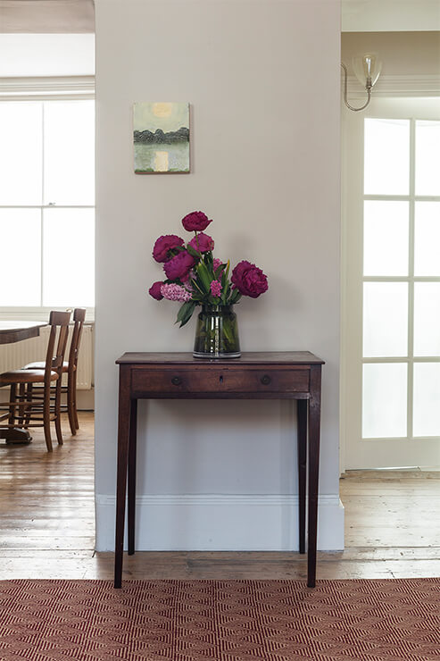 Floral arrangement in a vase on a wooden side table in a room with large windows.