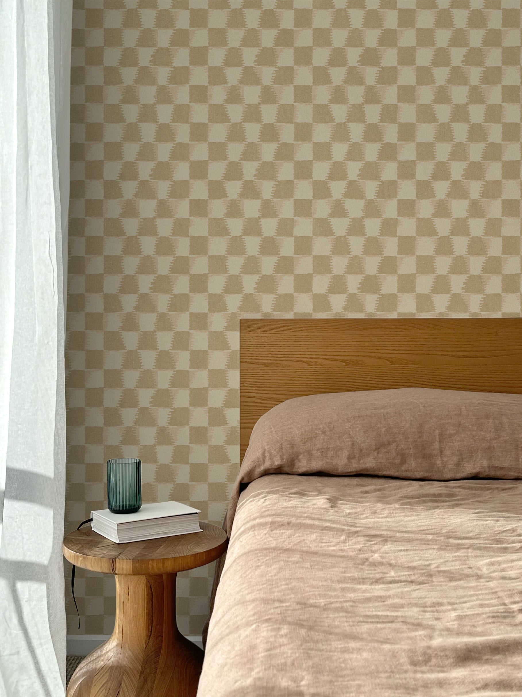 Bed with tan linens and wooden headboard, wooden side table  with white book and green glass, white floor length curtain against a watercolor checkerboard wallpaper in tonal warm tan. 