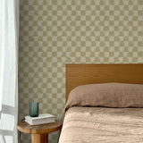 Bed with tan linens and wooden headboard, wooden side table  with white book and green glass, white floor length curtain against a watercolor checkerboard wallpaper in tonal sage green.