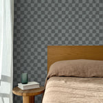 Bed with tan linens and wooden headboard, wooden side table  with white book and green glass, white floor length curtain against a watercolor checkerboard wallpaper in tonal cool charcoal grey. 