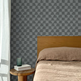 Bed with tan linens and wooden headboard, wooden side table  with white book and green glass, white floor length curtain against a watercolor checkerboard wallpaper in tonal cool charcoal grey. 