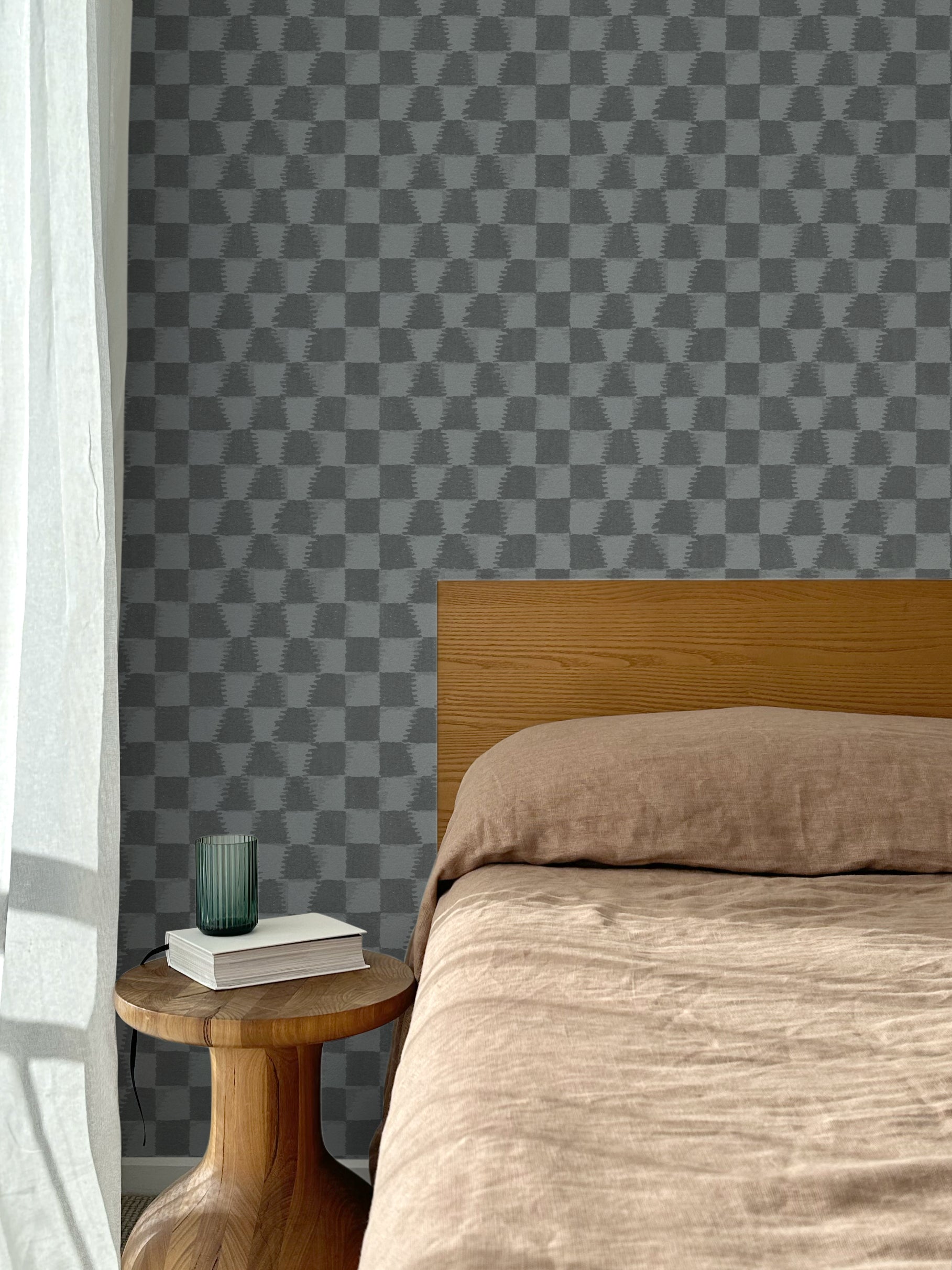 Bed with tan linens and wooden headboard, wooden side table  with white book and green glass, white floor length curtain against a watercolor checkerboard wallpaper in tonal cool charcoal grey. 