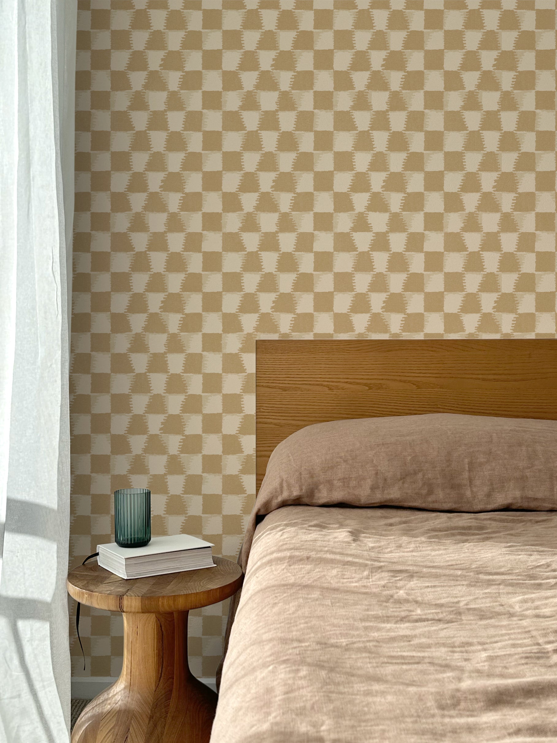 Bed with tan linens and wooden headboard, wooden side table  with white book and green glass, white floor length curtain against a watercolor checkerboard wallpaper in tan and beige.
