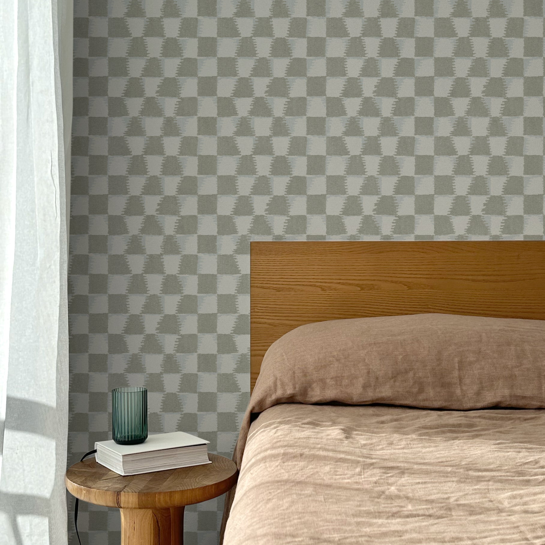 Bed with tan linens and wooden headboard, wooden side table  with white book and green glass, white floor length curtain against a watercolor checkerboard wallpaper in taupe-grey. 