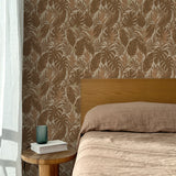 Bed with tan linens and wooden headboard, wooden side table  with white book and green glass, white floor length curtain against a brown tropical wallpaper. 